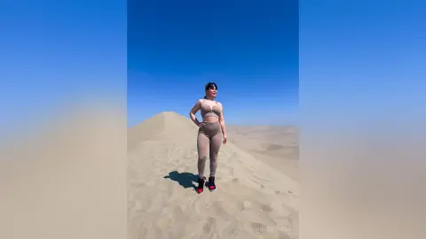 Woman in beige athletic wear standing on a vast sand dune under a clear blue sky.