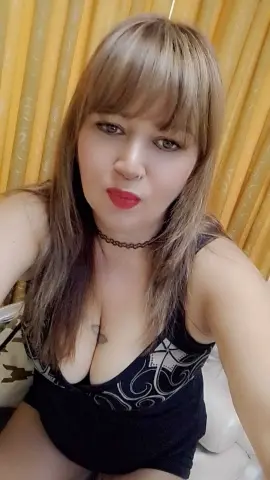 Young woman with blonde bangs in a black tank top posing against yellow curtains in a cozy indoor setting.