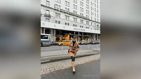 Stylish woman in a tan trench coat and cap walking on a city street near The Plaza Hotel in New York.