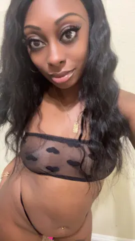 Smiling Black woman with long wavy hair wearing a black sheer polka dot bralette in a cozy indoor setting.
