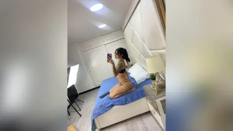 Tattooed woman in black lingerie sitting on a blue bed in a bright, modern bedroom taking a mirror selfie.