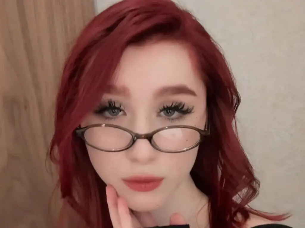 Close-up portrait of a young woman with red hair and glasses looking into the camera with a soft expression.