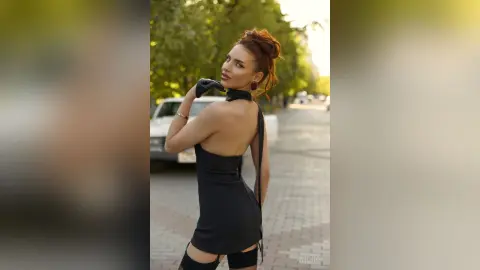 AnjeNoir poses on a city street wearing a tight black mini dress and black gloves, looking back over her shoulder.