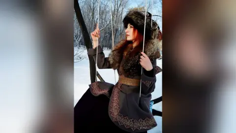 Red-haired woman in a vintage fur-trimmed coat sitting on a swing in a snowy winter forest.