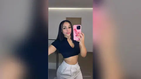 Young woman in a black crop top and white trousers taking a selfie in a modern minimalist room.