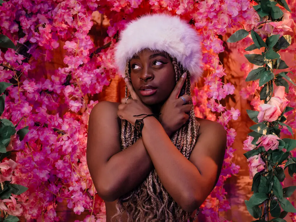 Dreamy portrait of a woman with long braids and a white faux fur hat amidst vibrant pink flowers.