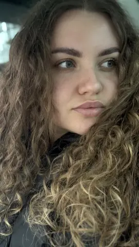 Close-up portrait of a young woman with long curly blonde hair wearing a black jacket in soft natural light.