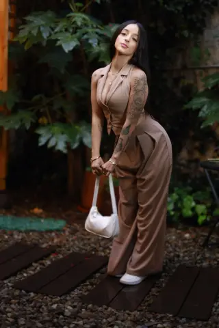 Young woman in a beige jumpsuit holding a white handbag in a lush garden setting.