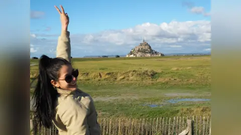 JessieLiu smiles and holds up a peace sign while standing in a grassy field under a blue sky.