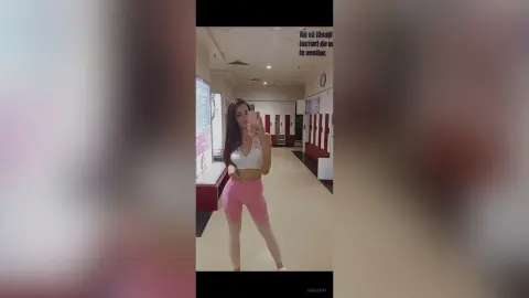 Young woman in white sports bra and pink leggings taking a mirror selfie in a modern gym hallway.
