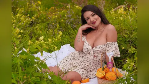 Smiling woman in a floral sundress sitting on a white blanket in a lush green meadow with a basket of oranges.