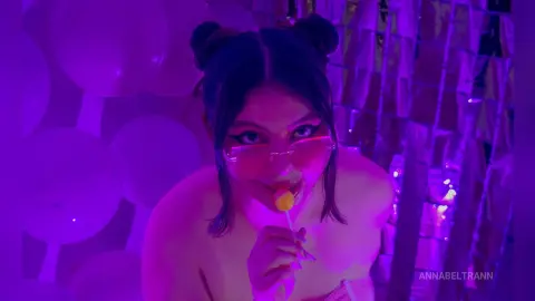AnnaBeltrann licks a yellow lollipop while wearing pink glasses and pink hair buns, set against a background of white balloons and purple lights.