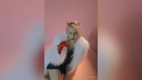 Glamorous blonde woman in a white faux fur coat and red gloves posing against a pink background.