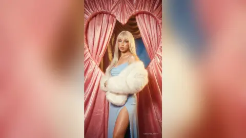 Glamorous blonde woman in a blue slit dress and white faux fur stole posing against pink heart-shaped curtains.