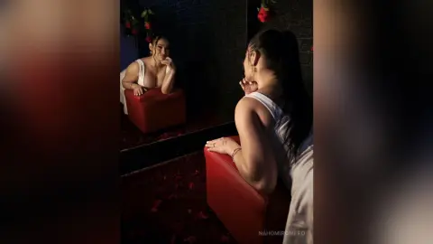 NahomiRomero poses in a white tank top, leaning over a red chair while looking back over her shoulder in a dark room.