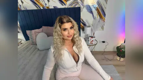 Blonde woman in white loungewear sitting on a bed in a cozy, modern bedroom setting.