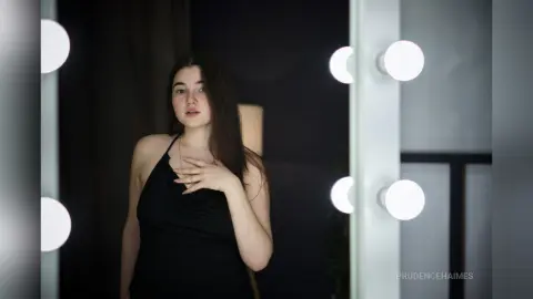 Young woman in a black halter top posing in a dimly lit room with vanity mirror lights.