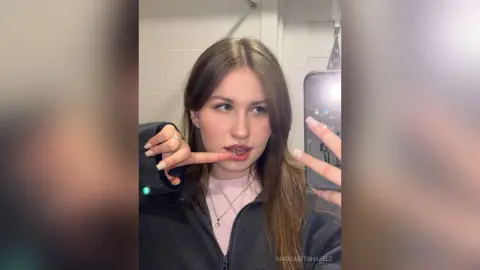 MargaritaHafele poses for a mirror selfie wearing a dark grey zip-up jacket and a pink top, running a finger along her bottom lip.