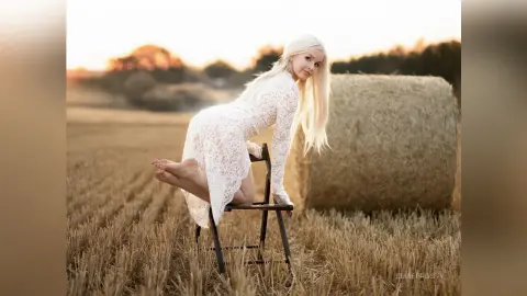 Blonde woman in a white lace dress sitting on a chair in a golden wheat field at sunset.