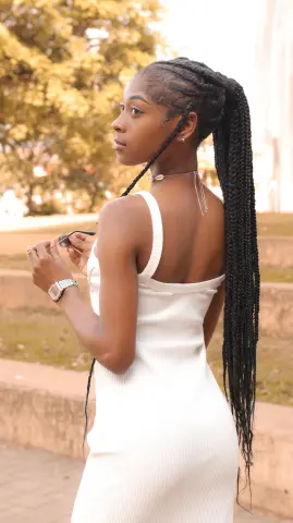 Young Black woman in a white ribbed dress with long braids posing outdoors in warm sunlight.