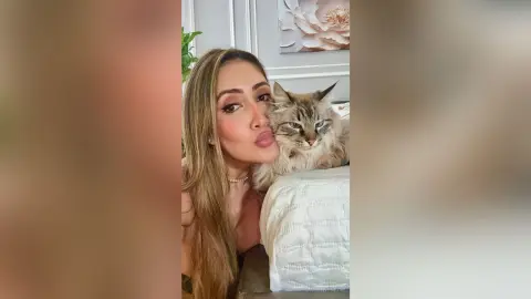 Smiling blonde woman cuddling a fluffy grey tabby cat in a bright, cozy indoor setting.