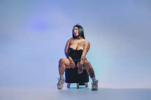 Tattooed woman in a black corset sitting on a stool against a blue studio background with moody lighting.