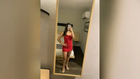 NatiSweet poses in a mirror wearing a short red dress and clear heels, standing in a bedroom with a bed and air conditioner visible behind her.