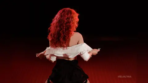 Red-haired woman in a white off-the-shoulder top and black skirt posing against a dark red studio background.