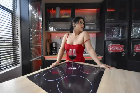 Elegant woman in a red dress leaning on a kitchen counter with a glass of red wine in a modern home setting.