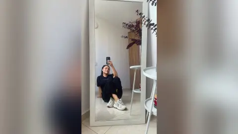 Young woman in black t-shirt and trousers taking a mirror selfie in a bright, minimalist modern room.