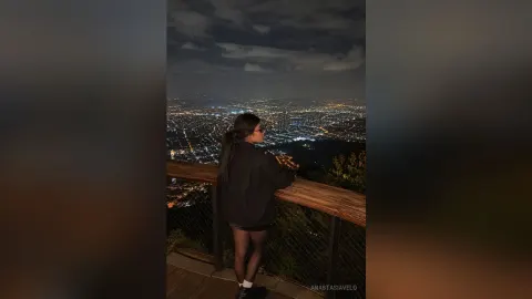 AnastasiaVelo — AnastasiaVelo stands on a balcony at night overlooking a bright city skyline while wearing a black long-sleeve shirt and shorts.