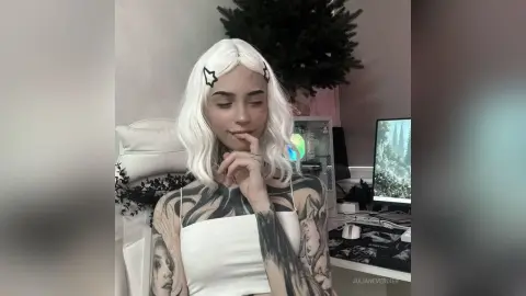 Young woman with white wavy hair and tattoos wearing a white tank top in a cozy, modern room.