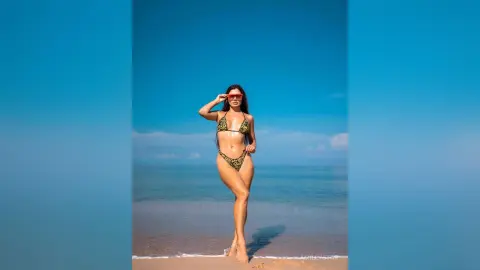 Young woman in a leopard print bikini posing on a sunny tropical beach under a clear blue sky.