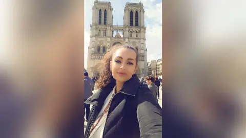 Smiling woman in a black puffer jacket taking a selfie in front of the Notre Dame Cathedral in Paris.
