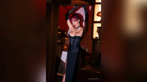 LysaraReyes poses with her hands behind her head, wearing a black lace corset and long black gloves against a dark red background.
