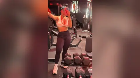 Fit woman in white sports bra and black leggings posing in a modern gym setting with dumbbells.