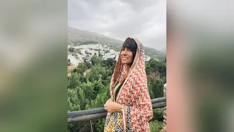 Smiling woman in a patterned traditional shawl overlooking a scenic mountain village under a cloudy sky.