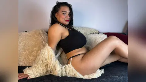 Smiling woman in a black crop top relaxing on a cozy white faux fur rug in a bright indoor setting.