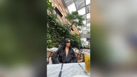 AriaBlaze sits at a marble table wearing black sunglasses and a black shirt, surrounded by lush green plants in a bright sunlit patio.