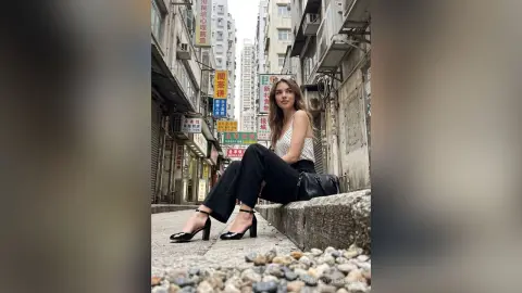 CharlotteReimer sits on a concrete ledge in a city alleyway, wearing a white tank top, black pants, and black high heels.
