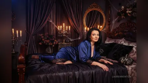 LaurenNewton — LaurenNewton lies on a black bed wearing a blue satin robe with lace trim in a dark, moody bedroom.