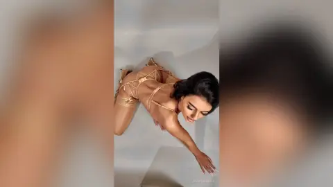 JasmineHille poses on all fours wearing a gold strappy lingerie set, looking up while showing her back and curves.