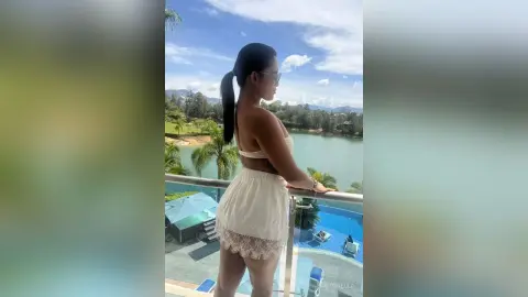 Woman in white lace bikini set relaxing on a balcony overlooking a tropical lake and mountains.