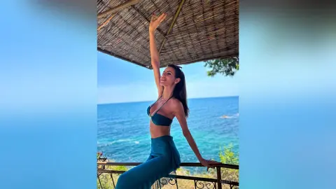 Smiling woman in a teal bikini top and trousers relaxing under a thatched roof by the blue ocean.