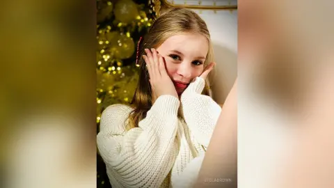 StelaBrown rests her chin on her hands while wearing a white knit sweater against a gold holiday background.