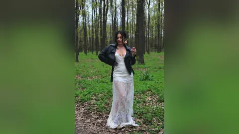Woman in a white lace wedding dress and black leather jacket standing in a lush green forest.