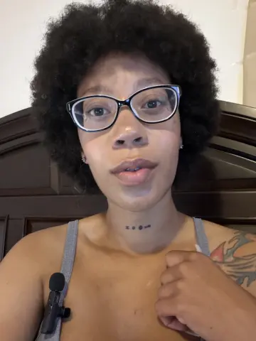 LexBright wearing black glasses and a grey tank top, showing her afro and neck tattoo while looking into the camera.