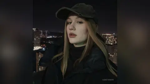 Young woman in a black jacket and baseball cap posing outdoors at night against a city skyline background.