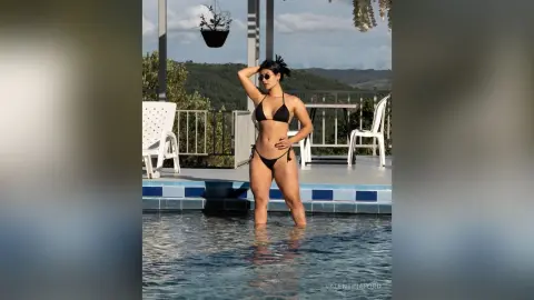 Confident woman in black bikini posing in a luxury swimming pool overlooking a scenic mountain landscape.