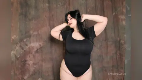 Curvy woman in a black bodysuit posing confidently against a textured brown studio backdrop.